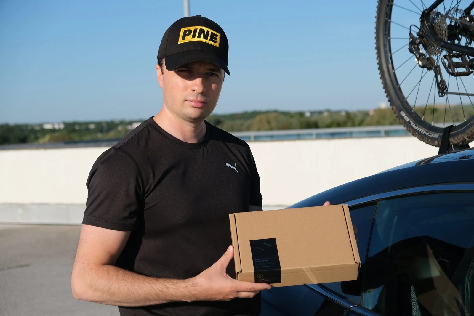 Timothy wearing a Pine cap, holding a Kraken Rack Monitor shipping box next to the car